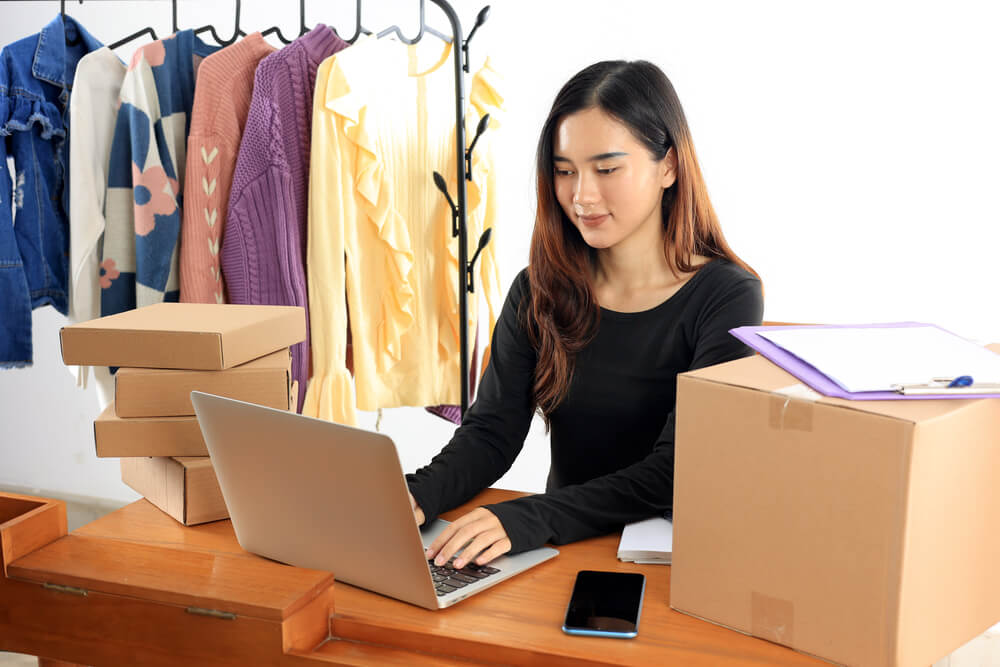 A woman on her laptop with boxes and clothes around her. Check here for the 12 ways single moms can build wealth in 2024.