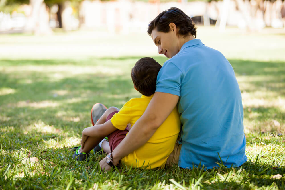 A man sitting on the ground with a child, his one arm over the child's shoulder. Check these 31 co-parenting tips to make shared custody a success.