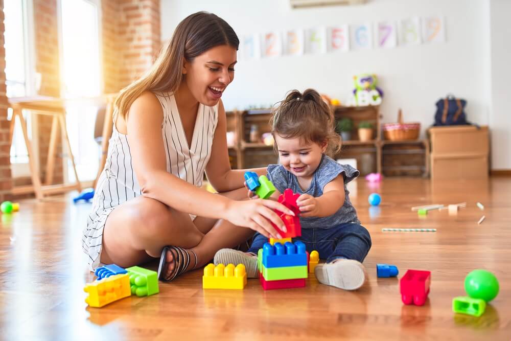 A woman playing blocks with a child on the floor. Find out about these 6 places to find a babysitter in 2024.