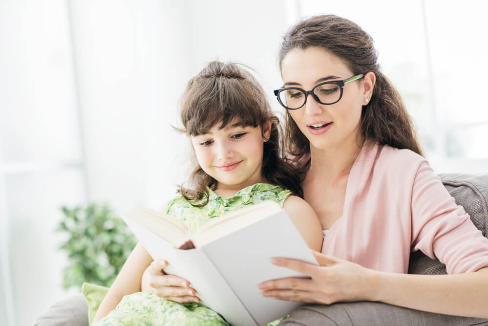 A bespectacled woman reading to a child. Check out these 6 places to find a babysitter in 2024 here.