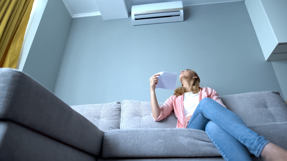 The woman is fanning herself while looking at the air conditioning unit. Some people struggle to meet energy bill expenses, but there is help available through assistance programs.