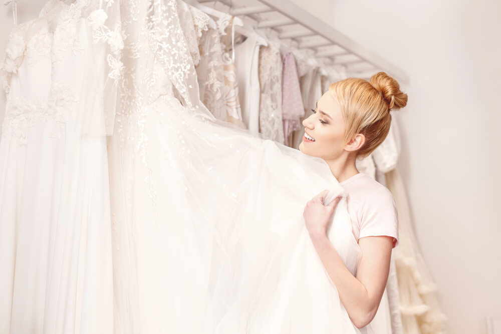 The woman smiles as she picks her wedding dress. You can pick yours, too online. Check sites like PreOwned WeddingDresses.com.