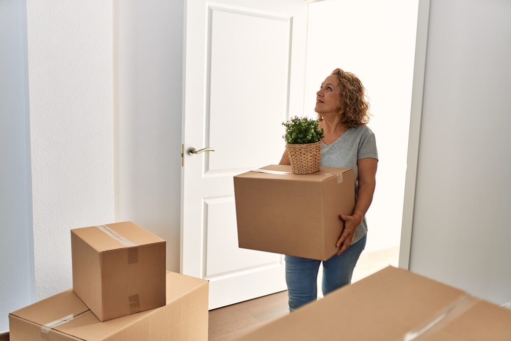 The middle-aged woman holds a cardboard box with a plant on top of the box as she walks into a new home. Fannie Mae offers mortgages with low down payments to make it easier for single moms to buy a home.