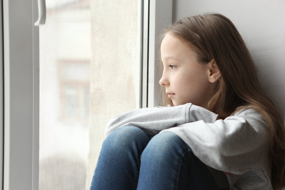 A lonely child looking out the window. What is parental alienation? Recognize the 18 signs you can find here.